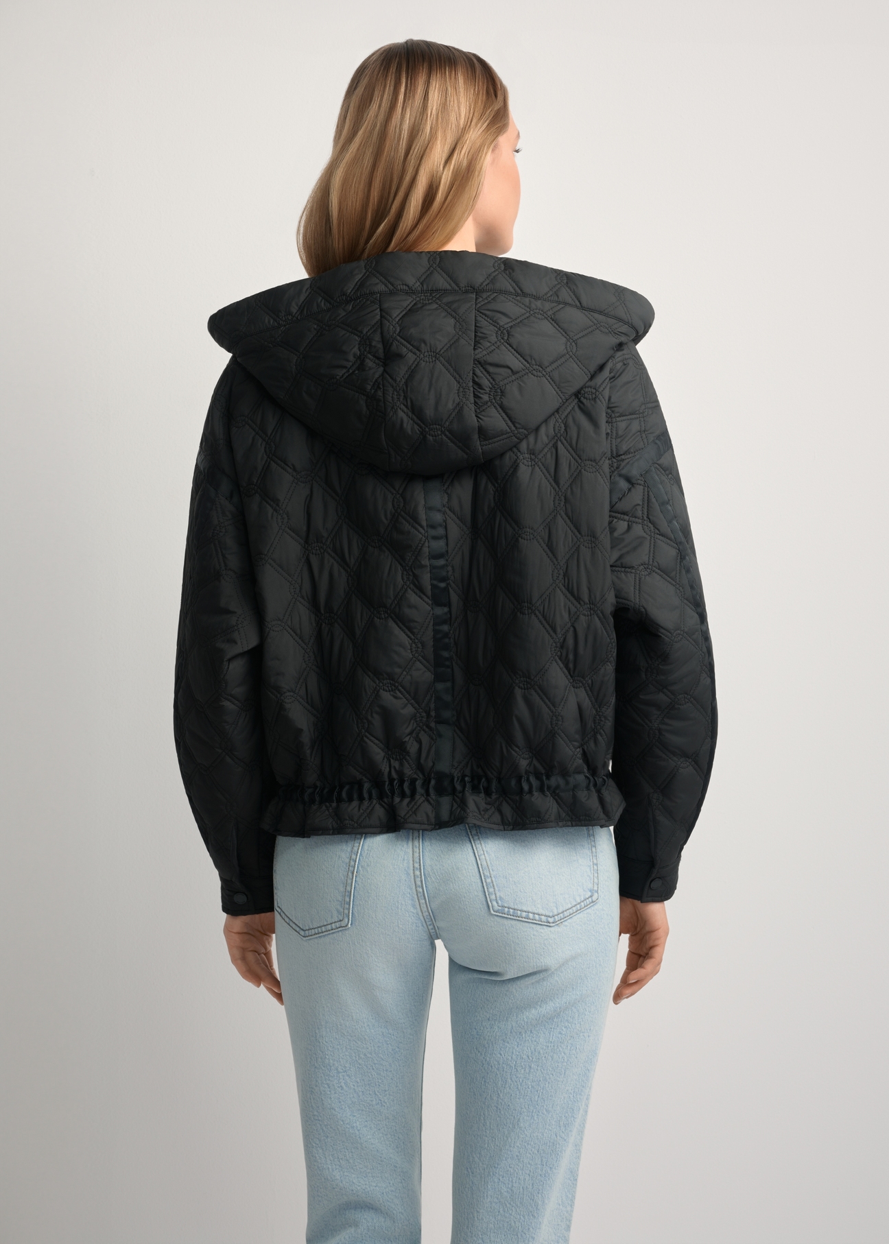 Czarna krótka pikowana kurtka oversize KURDT-0660-99(W26) - widok produktu z tyłu, na modelce, na jasnym tle. Widoczna sylwetka do kolan. 