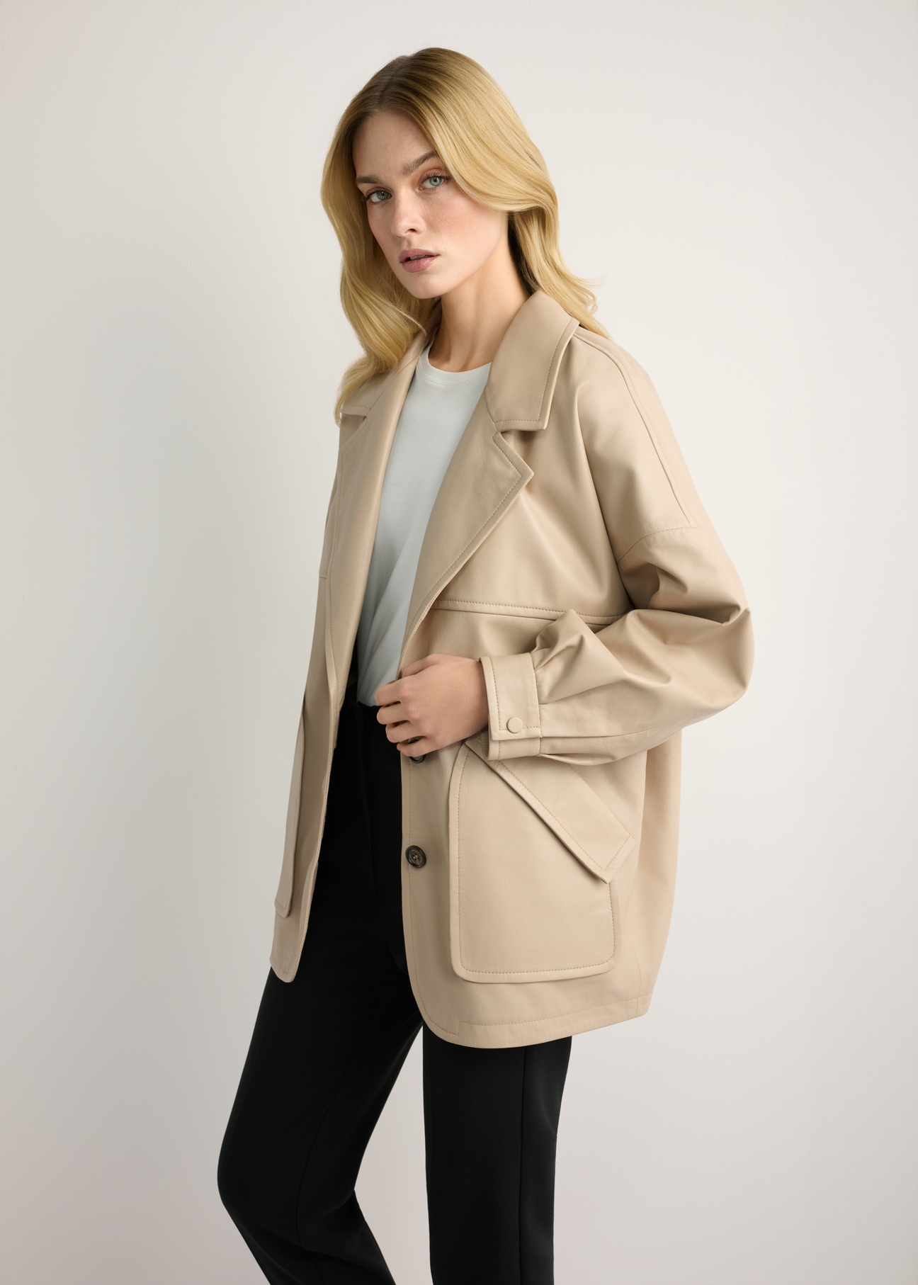 Beżowa skórzana kurtka damska oversize KURDS-0577-1B(W26) - widok produktu z boku, na modelce, na jasnym tle. Widoczna sylwetka do kolan. 