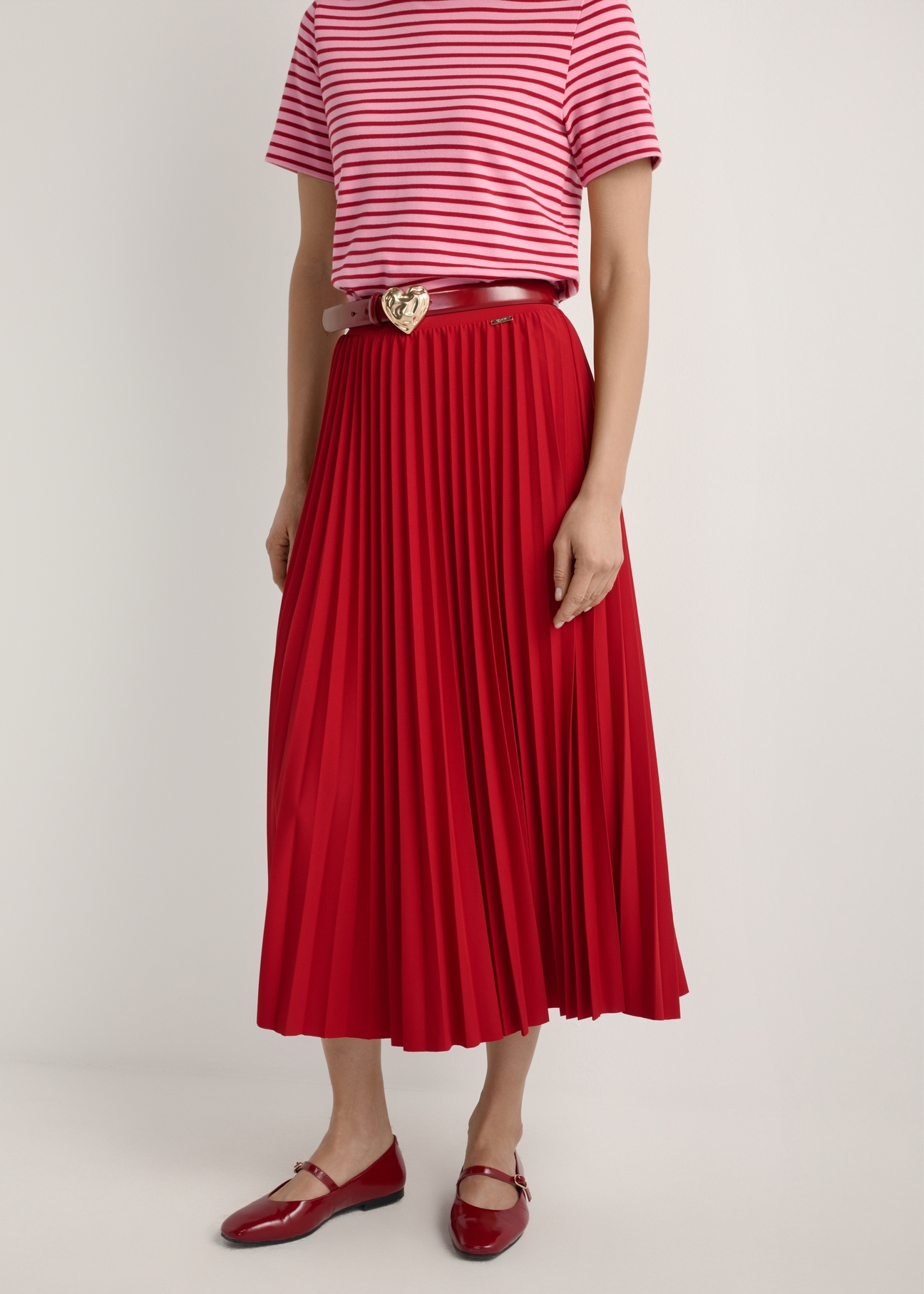 Czerwona plisowana spódnica midi SPCDT-0096-4B(W26), widok produktu od przodu na modelce, widoczna połowa sylwetki.