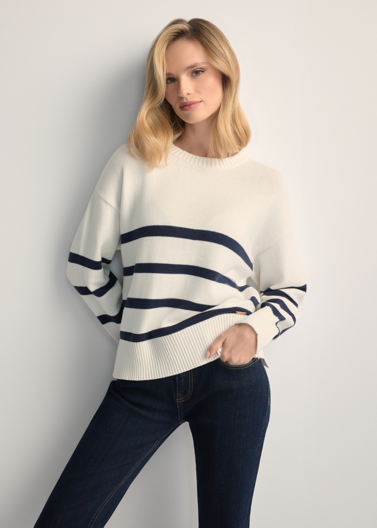 Kremowy sweter damski w paski SWEDT-0258-0B(W26) - widok produktu z przodu, na modelce, na jasnym tle. Widoczna sylwetka do kolan. 