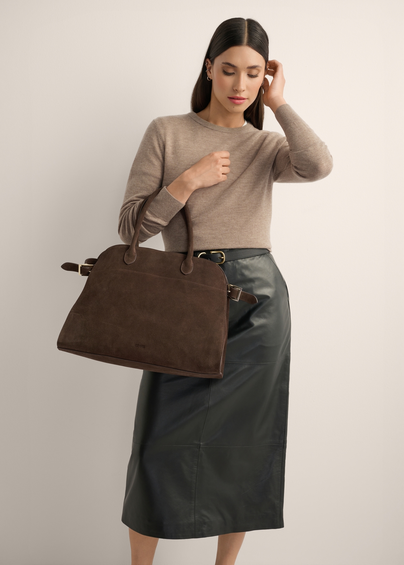 Ciemnobrązowa skórzana torebka damska typu shopper TORES-1412-1H(Z26) - widok torebki lekko po skosie, trzymanej przez modelkę w ręku, na jasnym tle. Widoczna sylwetka do kolan. 